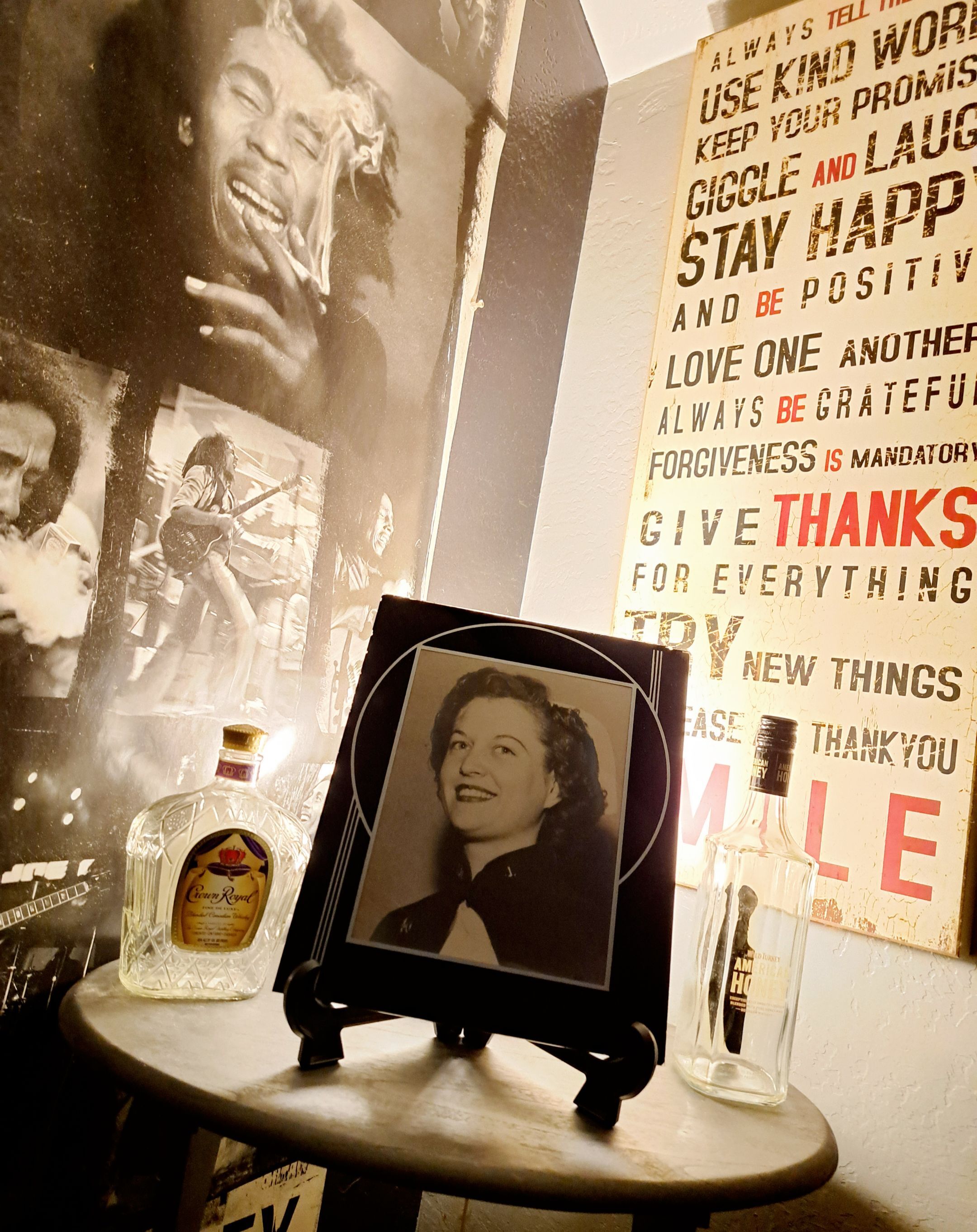 Sepia photo of a smiling woman on a round table with two liquor bottles. Background: black-and-white musician poster, motivational text. Cozy atmosphere.